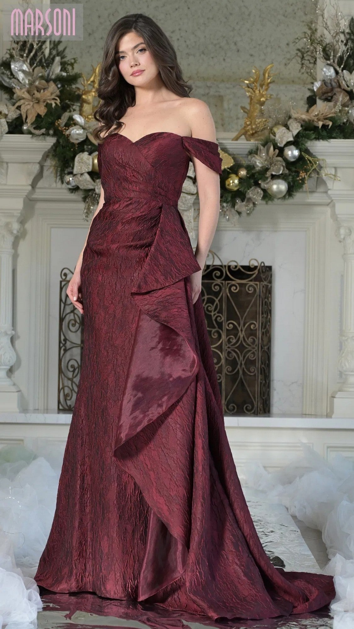 A woman wearing the Marsoni MV1365 long formal dress in burgundy stands indoors before a white fireplace decorated with ornaments and greenery, her elegant gown enhancing the festive atmosphere.